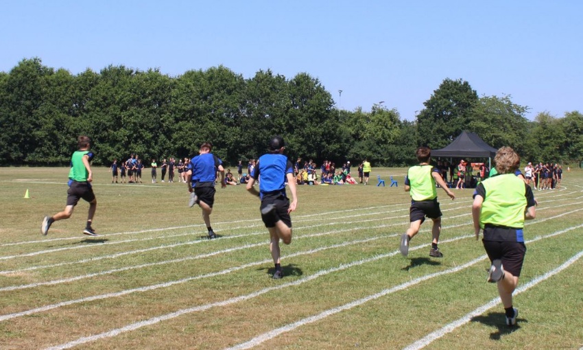 Pupils sprint along the running track during a relay race at Sports Day, with spectators and staff gathered beside the field.